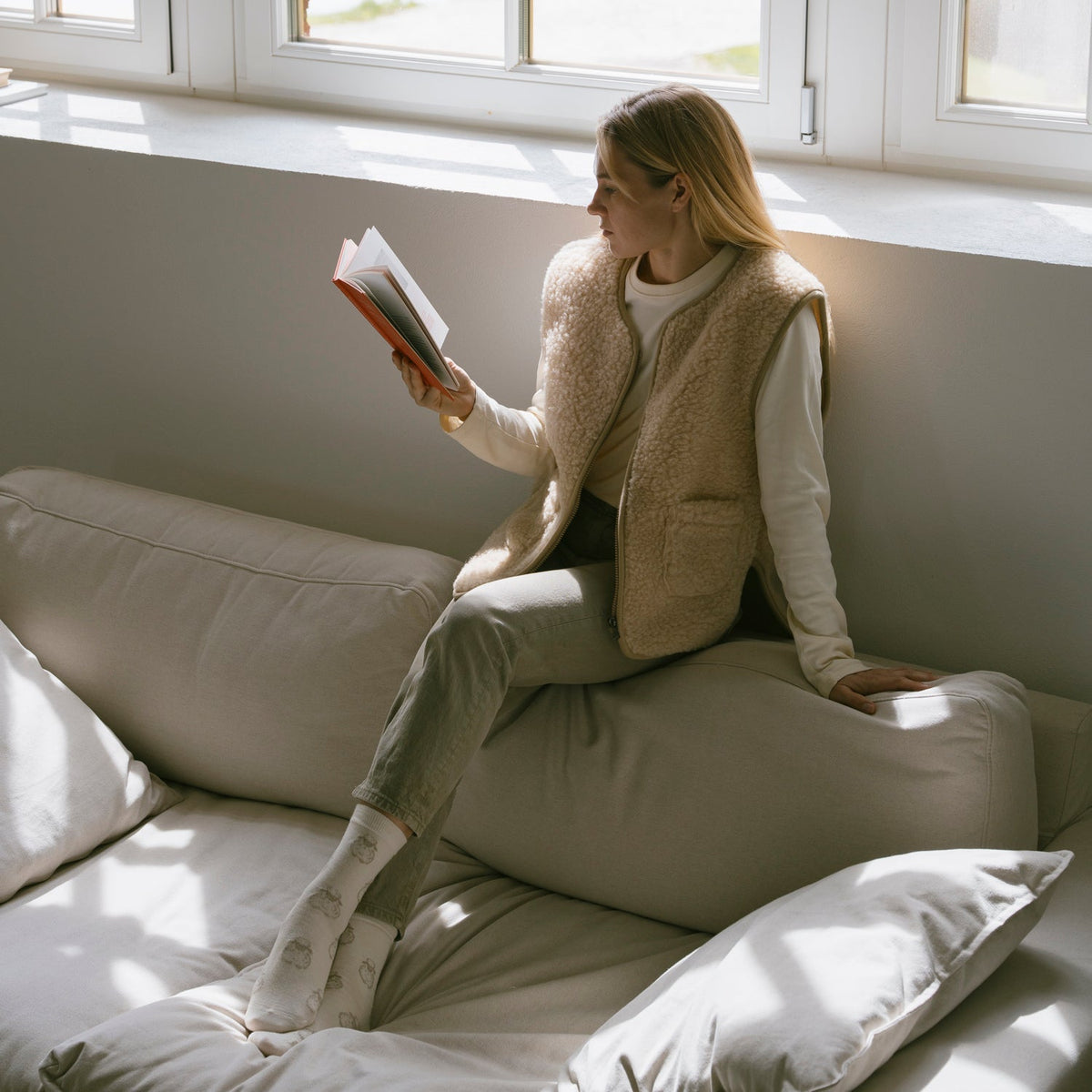 A woman reading on a sofa in the beige Island merino vest
