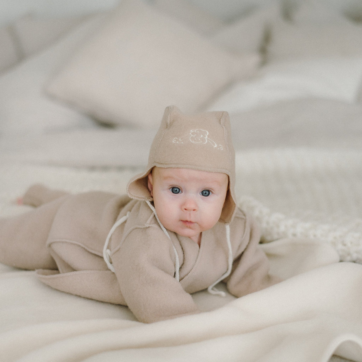 A baby lying on a blanket in the beige merino hat and jacket